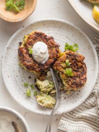 Small plate with two zucchini fritters, one cut into with a fork, the other topped with a dollop of sour cream.