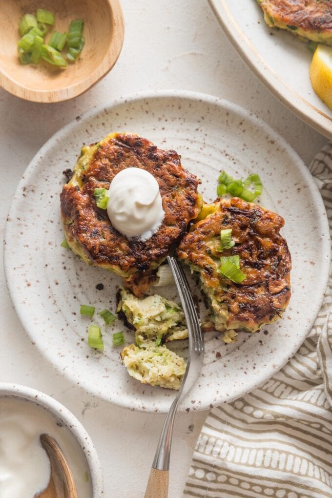 Small plate with two zucchini fritters, one cut into with a fork, the other topped with a dollop of sour cream.