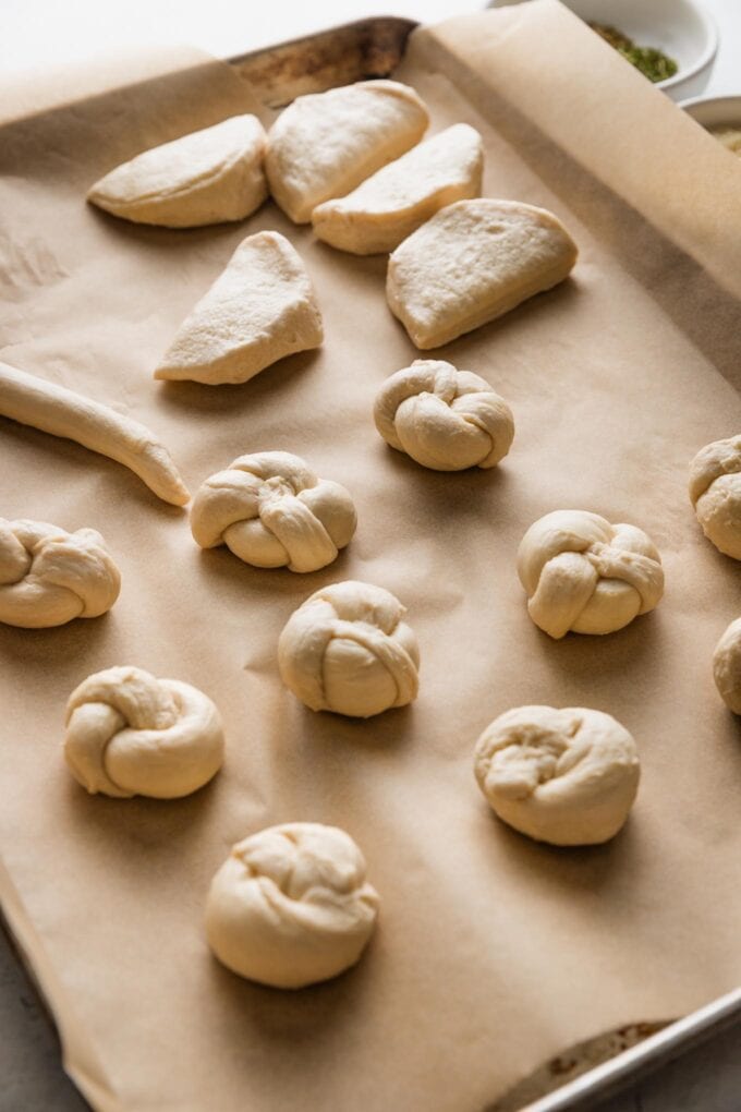 Large sheet pan lined with parchment paper and filled with garlic knot dough in various stages of tying and twisting.