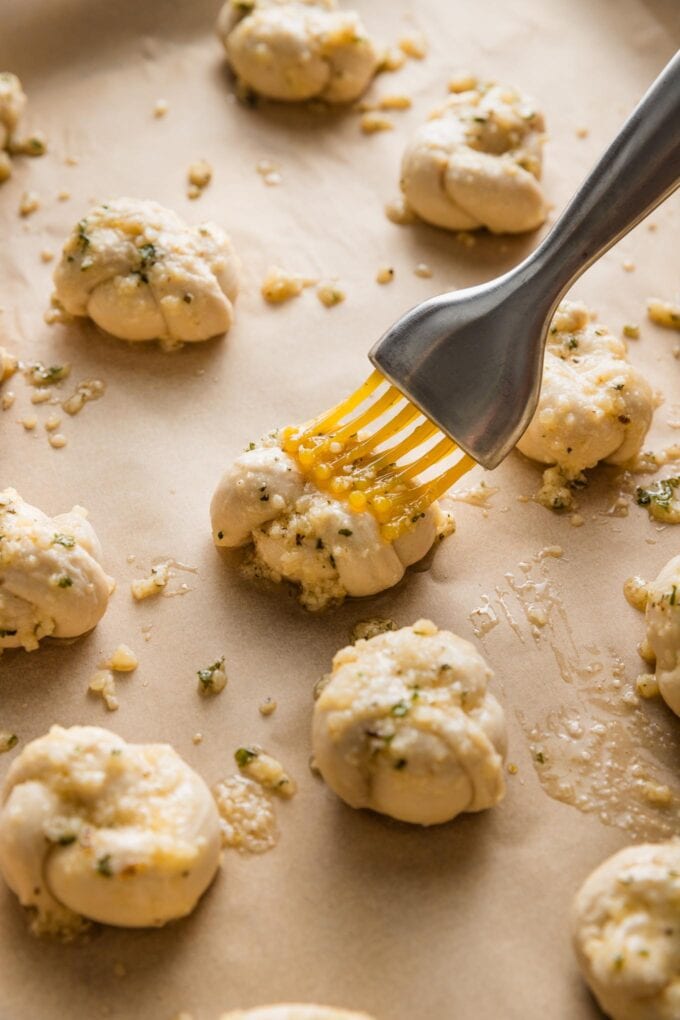 Close up view of a pastry brush coating small, unbaked garlic knots with a butter Parmesan herb mixture.