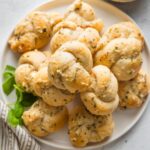 Small white plate piled high with garlic Parmesan biscuit garlic knots with fresh basil scattered nearby.