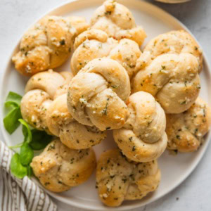 Small white plate piled high with garlic Parmesan biscuit garlic knots with fresh basil scattered nearby.