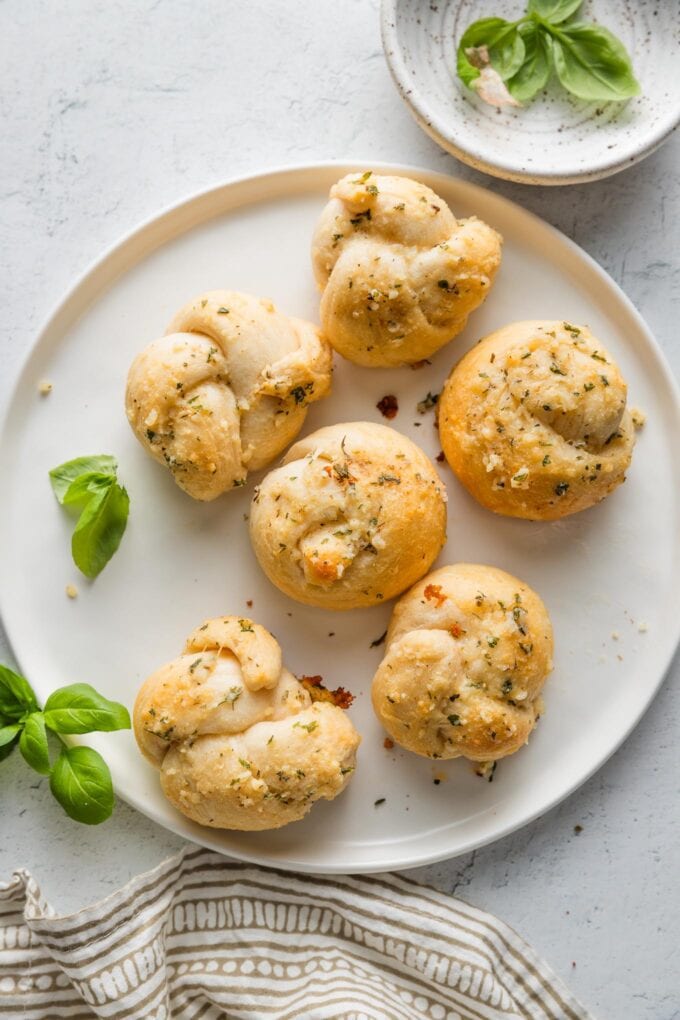 Countertop set with a small plate of garlic knots made out of biscuit dough, a patterned napkin, and extra basil leaves.