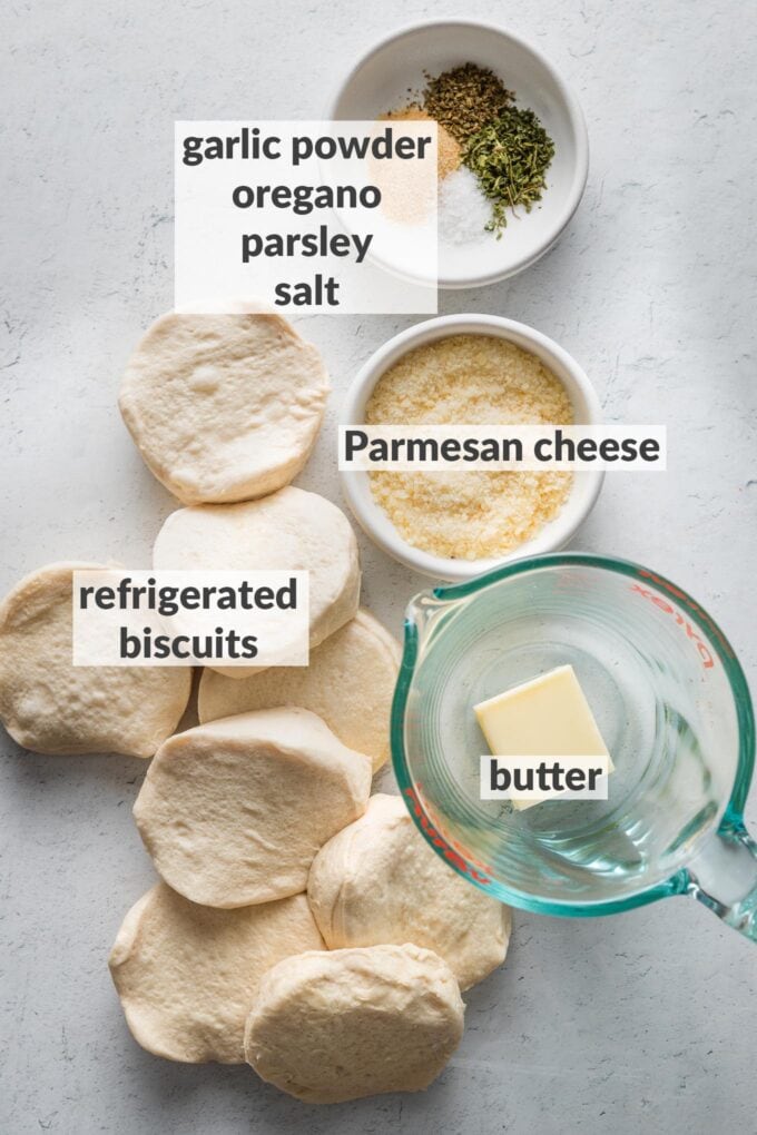 Labeled overhead image of refrigerated biscuit dough, butter in a liquid measuring cup, Parmesan cheese, garlic powder, parsley, dried oregano, and salt.