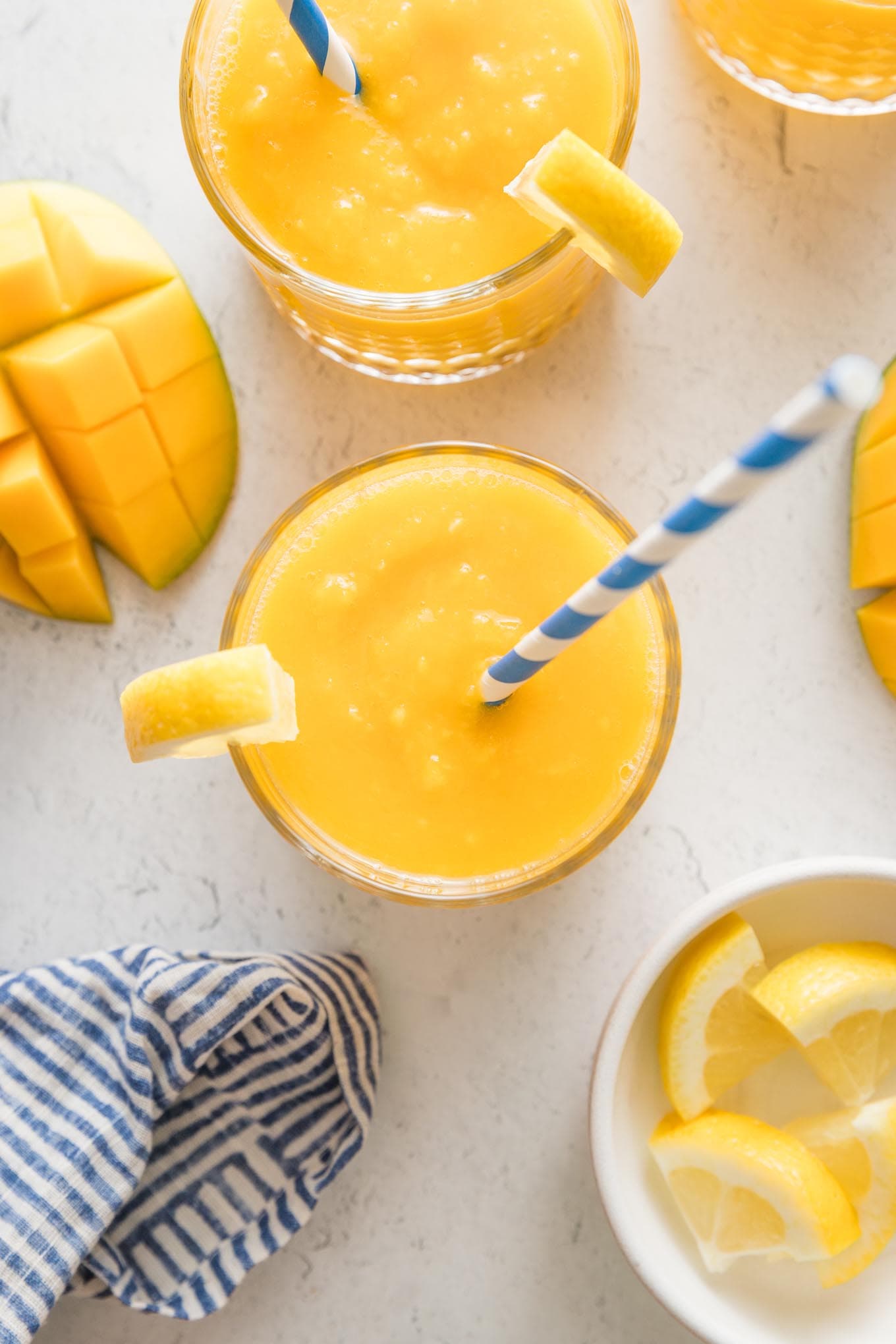 Overhead view of a small glass filled with frozen mango lemonade topped with a lemon wedge and a blue and white striped paper straw.