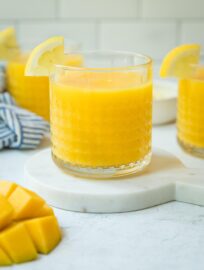 Countertop set with glasses of frozen mango lemonade plus extra lemon wedges and chunks of fresh mango.