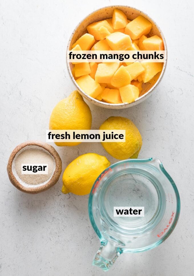 Labeled overhead photo of fresh lemons, cubed frozen mango chunks, two tablespoons of granulated sugar, and water in a liquid measuring cup.