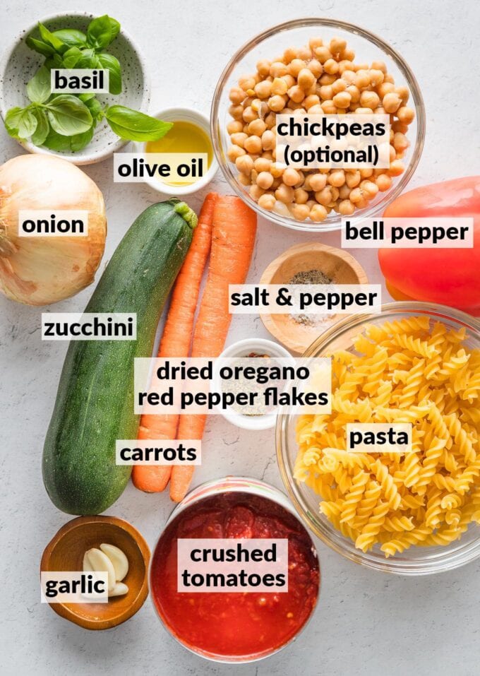 Labeled overhead photo of dried pasta, zucchini, carrots, crushed tomatoes, garlic, onion, bell pepper, chickpeas, fresh basil, olive oil, dried oregano, red pepper flakes, salt, and pepper, all measured into prep bowls and ready to cook.