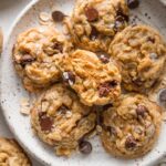 Close up view of mini oatmeal chocolate chip cookies with brown butter and sea salt, just out of the oven and stacked on a small ceramic plate with a cup of milk nearby.