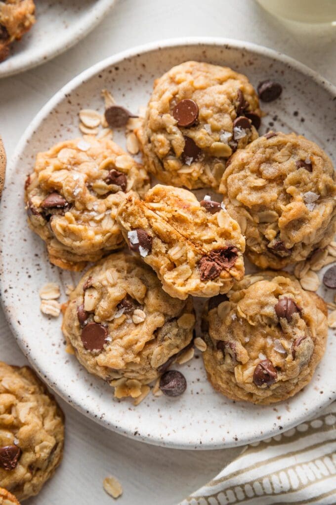 Close up view of mini oatmeal chocolate chip cookies with brown butter and sea salt, just out of the oven and stacked on a small ceramic plate with a cup of milk nearby.