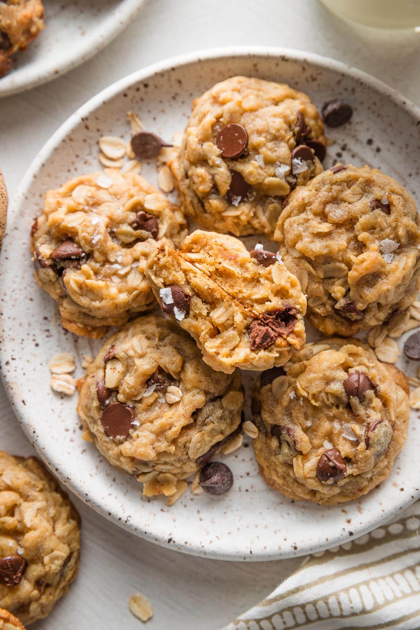 Close up view of mini oatmeal chocolate chip cookies with brown butter and sea salt, just out of the oven and stacked on a small ceramic plate with a cup of milk nearby.