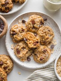 Small plate filled with mini oatmeal chocolate chip cookies with one split in half to show the tender, golden interior, with extra cookies and a glass of milk resting on the countertop nearby.