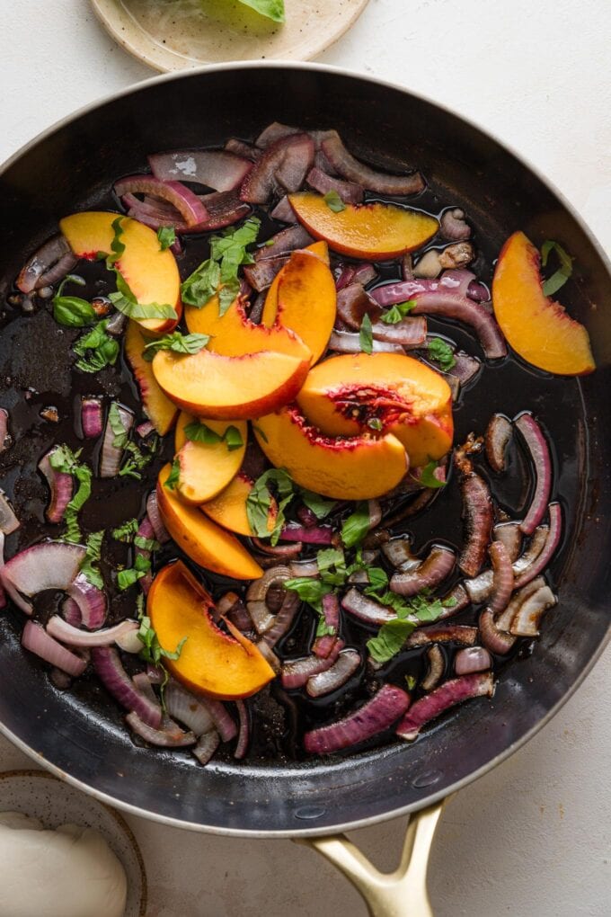 Large skillet filled with thinly-sliced red onions, fresh sliced peaches, and slivered basil.