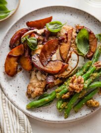 Speckled white ceramic dinner plate piled with a serving of balsamic peach mozzarella chicken and a side of pesto green beans with walnuts.