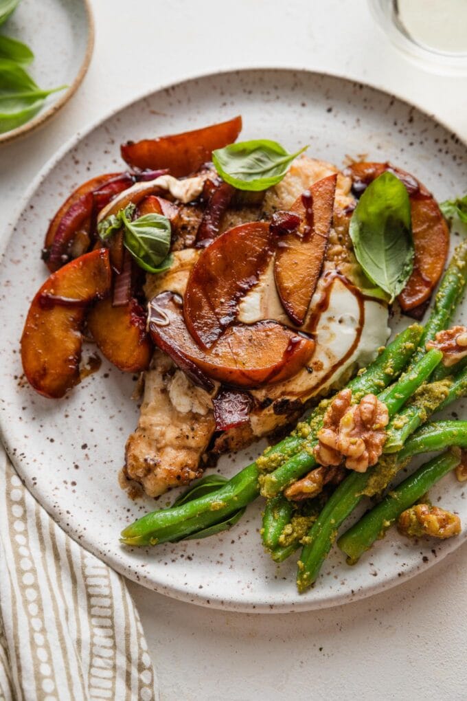 Speckled white ceramic dinner plate piled with a serving of balsamic peach mozzarella chicken and a side of pesto green beans with walnuts.