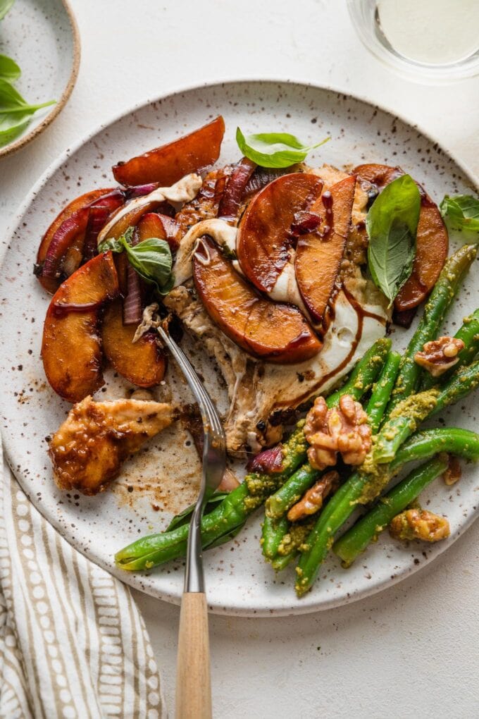 Small plate with a helping of peach mozzarella chicken with a fork lifting out a bite to show how tender it is, served with pesto green beans on the side.