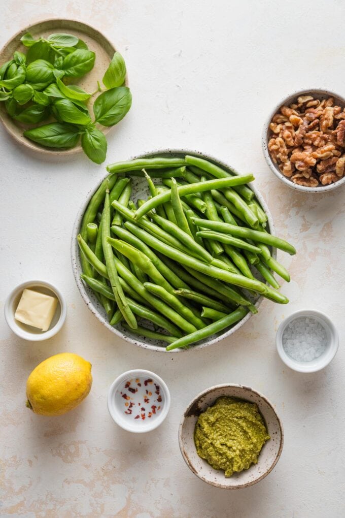Overhead photo of prep bowls containing green beans, fresh basil, pesto, walnuts, butter, a lemon, salt, and red pepper flakes.