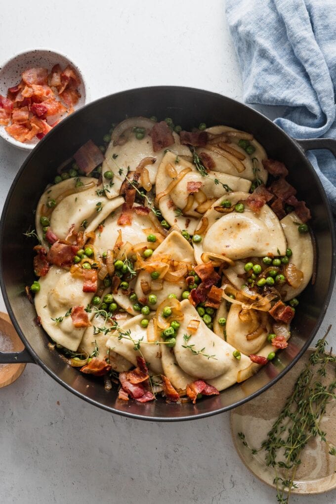 Overhead view of a large cast iron skillet filled with pierogies, caramelized onions, brown butter, peas, and bacon.