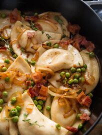 Close up of pierogies in a skillet with lightly browned butter, caramelized onions, peas, and bacon.