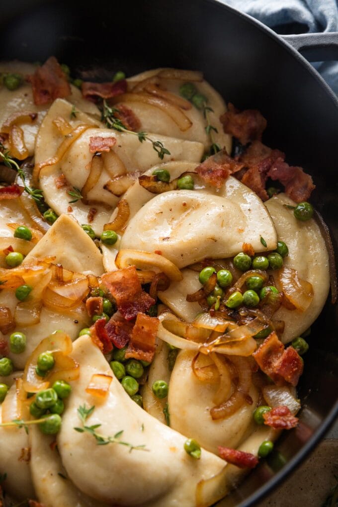 Close up of pierogies in a skillet with lightly browned butter, caramelized onions, peas, and bacon.