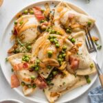 Small white plate filled with cheddar pierogies in a sauce of butter, caramelized onions, bacon, and peas.
