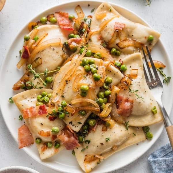 Small white plate filled with cheddar pierogies in a sauce of butter, caramelized onions, bacon, and peas.