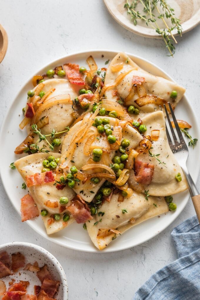 Small white plate filled with cheddar pierogies in a sauce of butter, caramelized onions, bacon, and peas.
