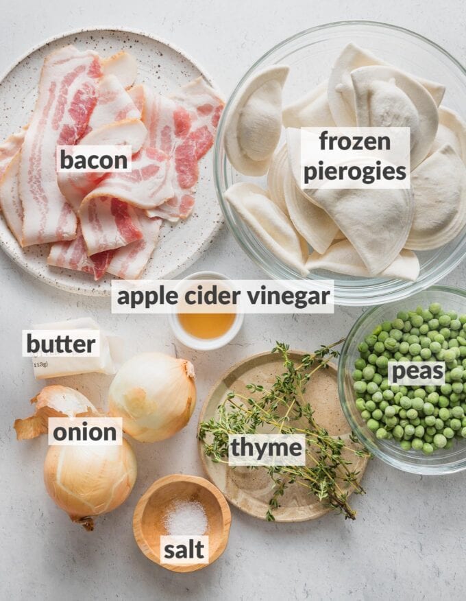 Labeled overhead photo of frozen pierogies, uncooked bacon slices, a whole yellow onion, frozen peas, fresh thyme, butter, apple cider vinegar, and salt, all measured into prep bowls and ready to cook.