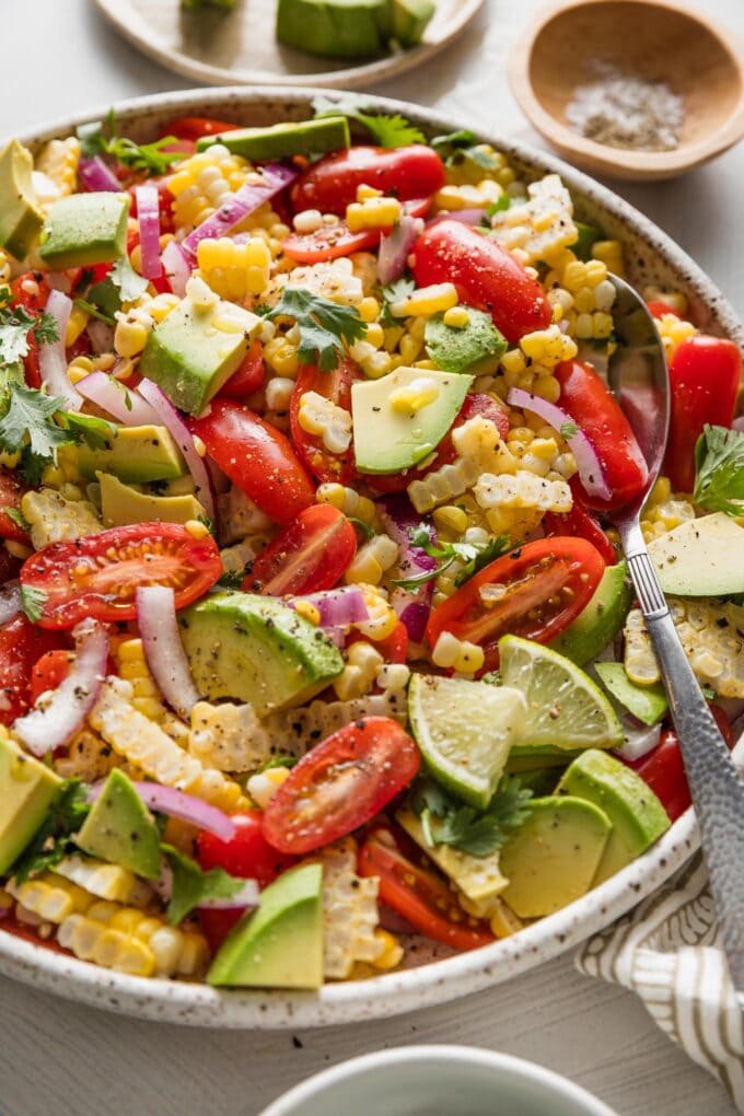 Angled view of a corn avocado tomato salad in a bowl with a large serving spoon.