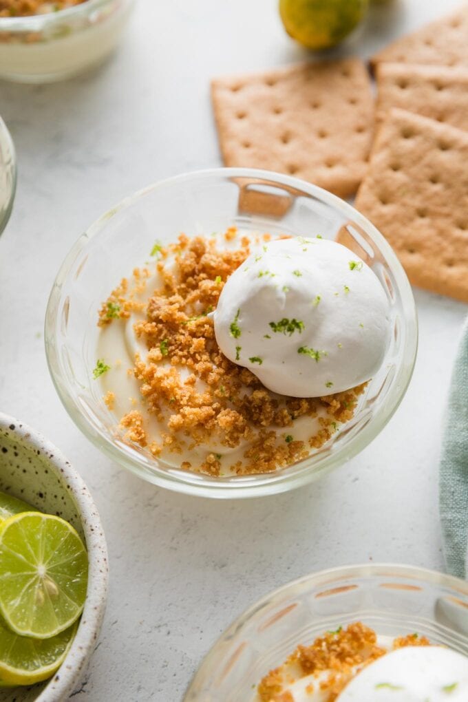 Small custard dish filled with a mini no bake key lime pie, served "upside down" with graham cracker crumbs, a dollop of whipped cream, and extra lime zest on top.