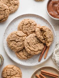 Speckled white plate piled with apple butter snickerdoodle cookies, with extra cookies, cinnamon sticks, and apple butter nearby on the counter.