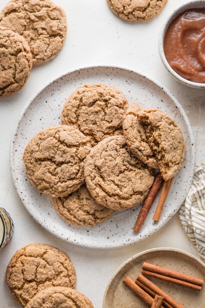 Speckled white plate piled with apple butter snickerdoodle cookies, with extra cookies, cinnamon sticks, and apple butter nearby on the counter.
