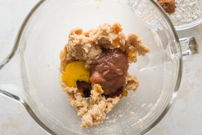 Overhead view of a clear glass stand mixer bowl holding creamed butter and sugar with an egg yolk, vanilla bean paste, and apple butter added but not yet mixed in.