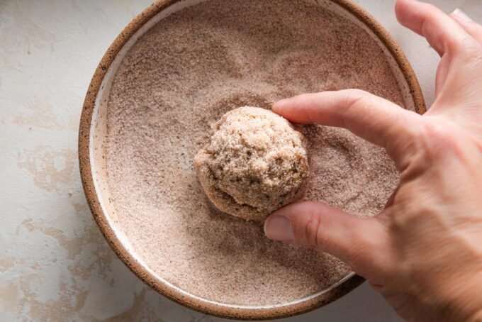 An action shot of someone rolling a snickerdoodle cookie dough ball in a small bowl of cinnamon sugar coating.