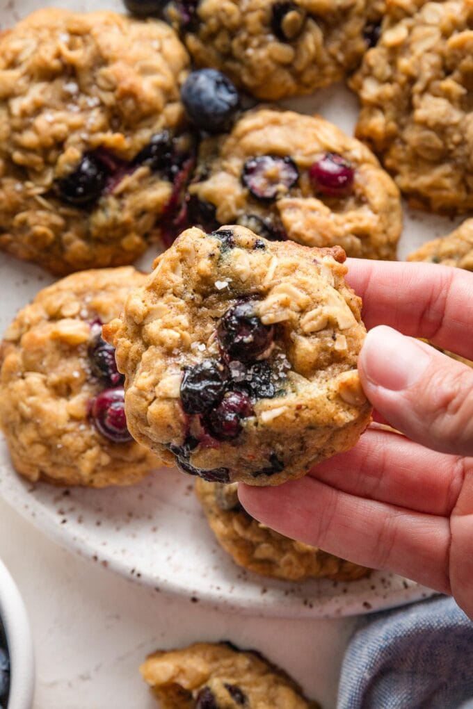 Hand holding up a small blueberry oatmeal cookie to show the soft texture of the cookie studded with juicy blueberries and nutty old-fashioned oats.