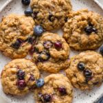 Close up of blueberry oatmeal cookies on a plate, with extra bowls of old fashioned oats and fresh blueberries scattered nearby.