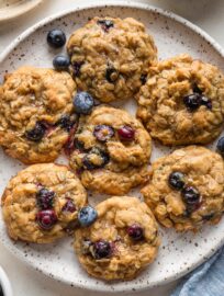 Close up of blueberry oatmeal cookies on a plate, with extra bowls of old fashioned oats and fresh blueberries scattered nearby.