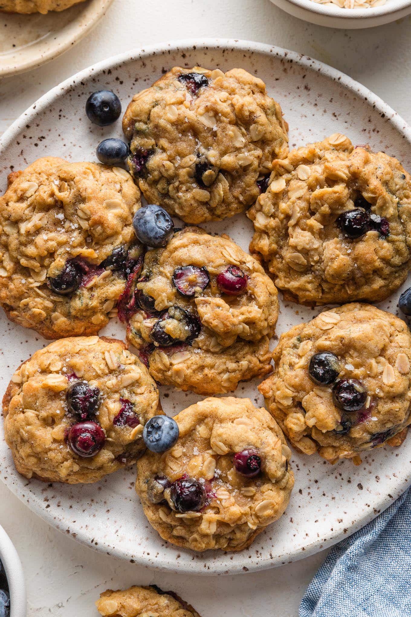 Close up of blueberry oatmeal cookies on a plate, with extra bowls of old fashioned oats and fresh blueberries scattered nearby.