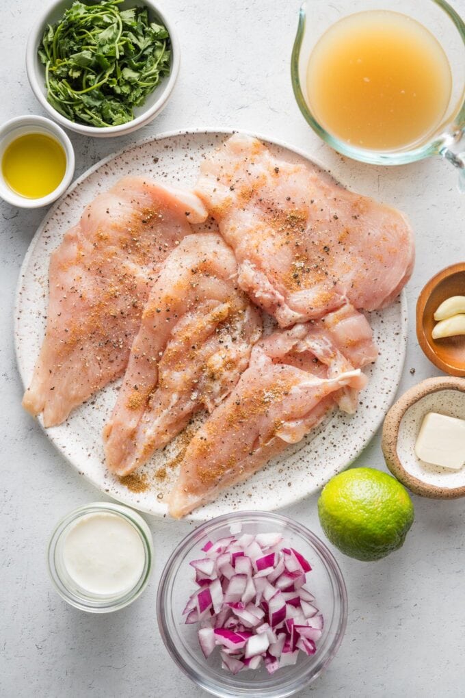 Overhead image of chicken breasts seasoned with salt, pepper, and cumin, chicken broth, cream, diced red onion, garlic cloves, a lime, butter, olive oil, and fresh cilantro leaves, all in prep bowls and ready to cook.