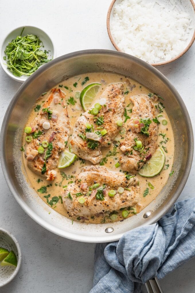 Overhead view of a large skillet filled with a dish of creamy cilantro lime chicken breasts, with extra cilantro, green onions, and white rice in small bowls on the countertop nearby.