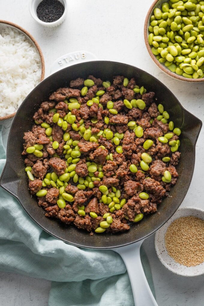 Countertop set with a skillet full of ground beef with edamame and separate bowls of jasmine rice, extra shelled edamame, and white and black sesame seeds for garnish.