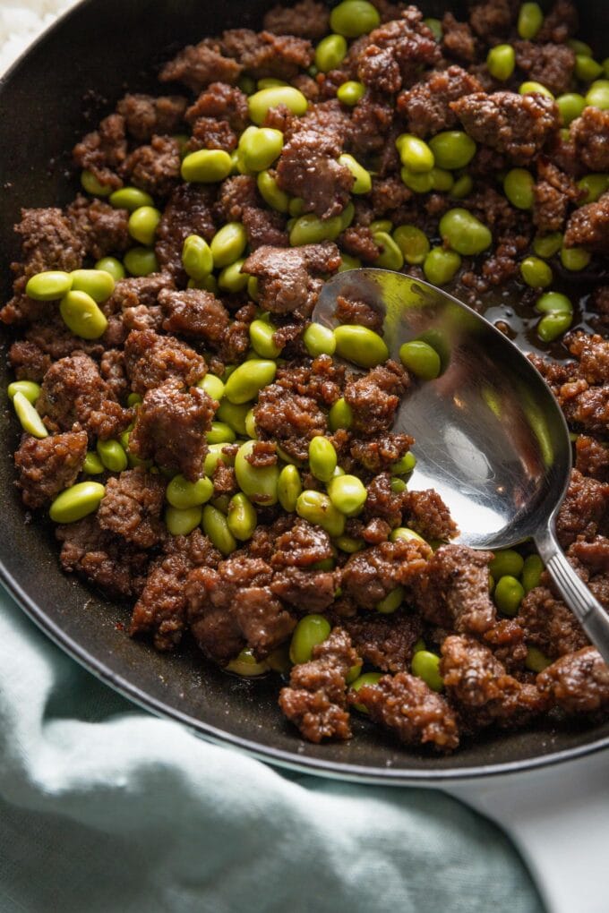 Close up of a serving spoon stirring a portion of ground beef with edamame in a small cast iron skillet.