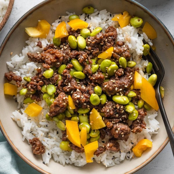 Ground beef bowl with edamame, yellow bell pepper, and sesame seeds served over white rice with a fork stirring everything together.