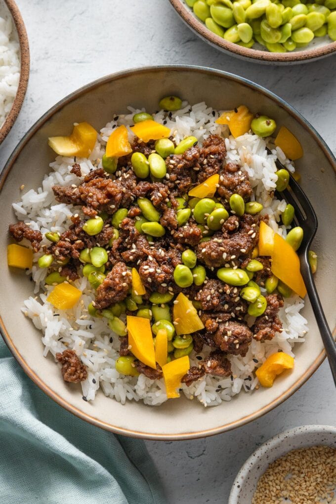 Ground beef bowl with edamame, yellow bell pepper, and sesame seeds served over white rice with a fork stirring everything together.