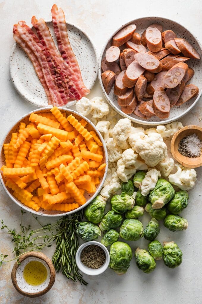 Overhead image of sliced chicken sausage, crisped bacon, Brussels sprouts, butternut squash crinkles, and cauliflower florets, with salt, pepper, olive oil, rosemary, and thyme for seasoning.