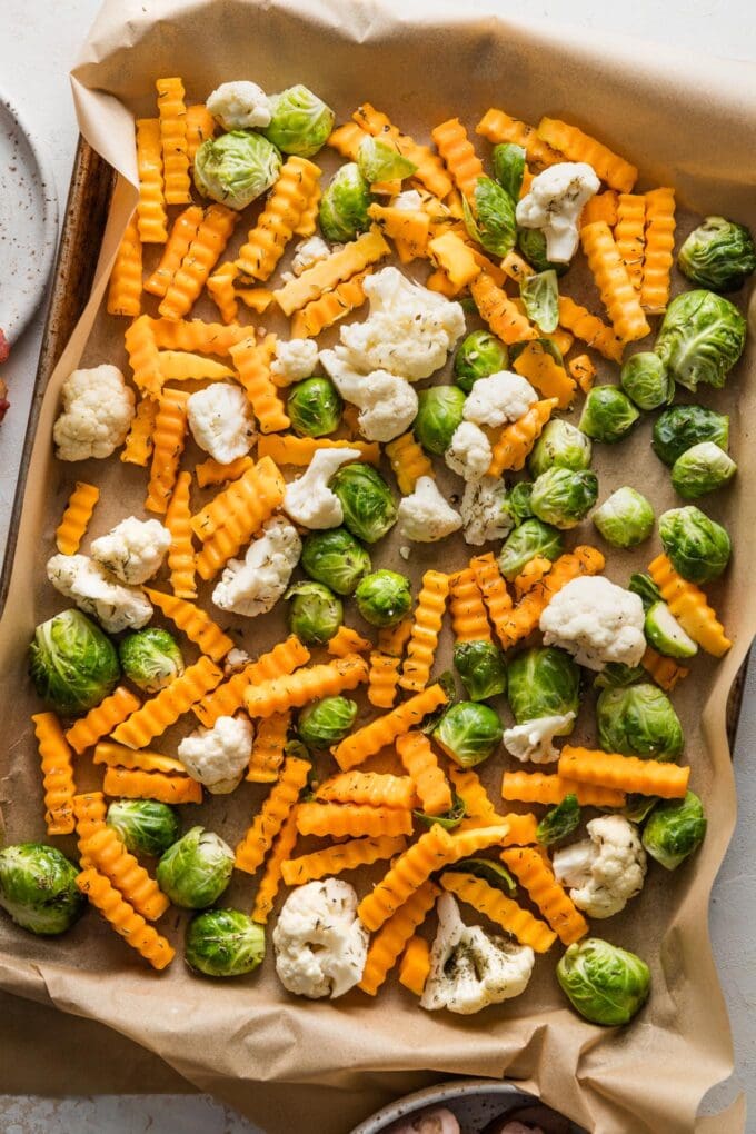 Halved Brussels sprouts, crinkle cut butternut squash, and cauliflower florets seasoned and spread out in a single layer on a parchment lined sheet pan.