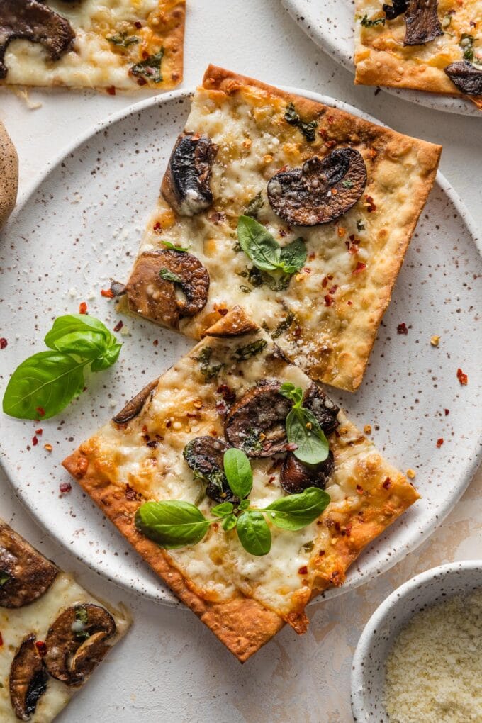 Small plate with two slices of a crisp mushroom flatbread pizza with mozzarella, fresh basil, and red pepper flakes.