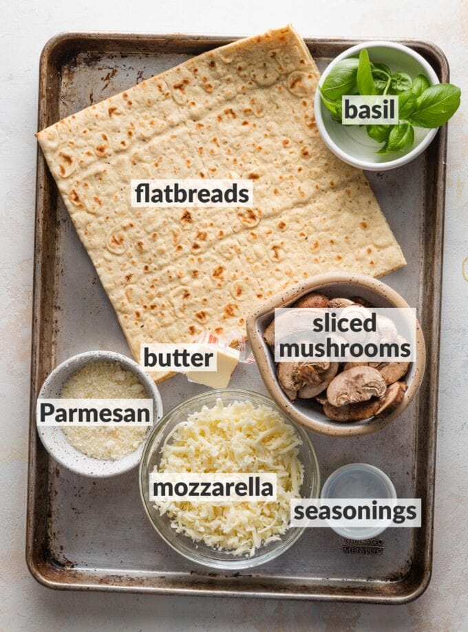 Large baking tray with lavash-style flatbread crusts, fresh basil, sliced mushrooms, butter, grated Parmesan cheese, and a small bowl of seasonings, all prepped and ready to assemble.