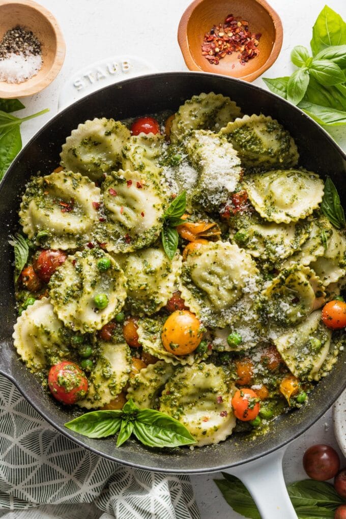 Cast iron skillet filled with a dish of pesto ravioli with peas and tomatoes, with extra basil scattered around for garnish.