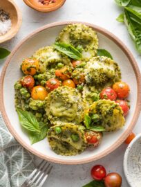 Small pasta bowl filled with pesto ravioli with peas and cherry tomatoes, with extra red pepper flakes nearby for garnish.